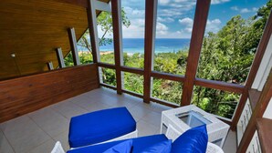 A balcony with two wicker chairs and blue cushions, overlooking a lush green landscape and the ocean. - Incredible Ocean views from both sides of this hill top home. (Roatan)