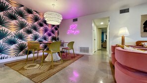 Een moderne woonkamer met een roze bank, een eethoek met gele stoelen, een neonbord en een behang met patroon.