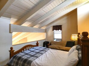 Un dormitorio con techo inclinado, ventana con un visillo marrón, cama con manta escocesa, lámpara de noche y cabecera de madera.
