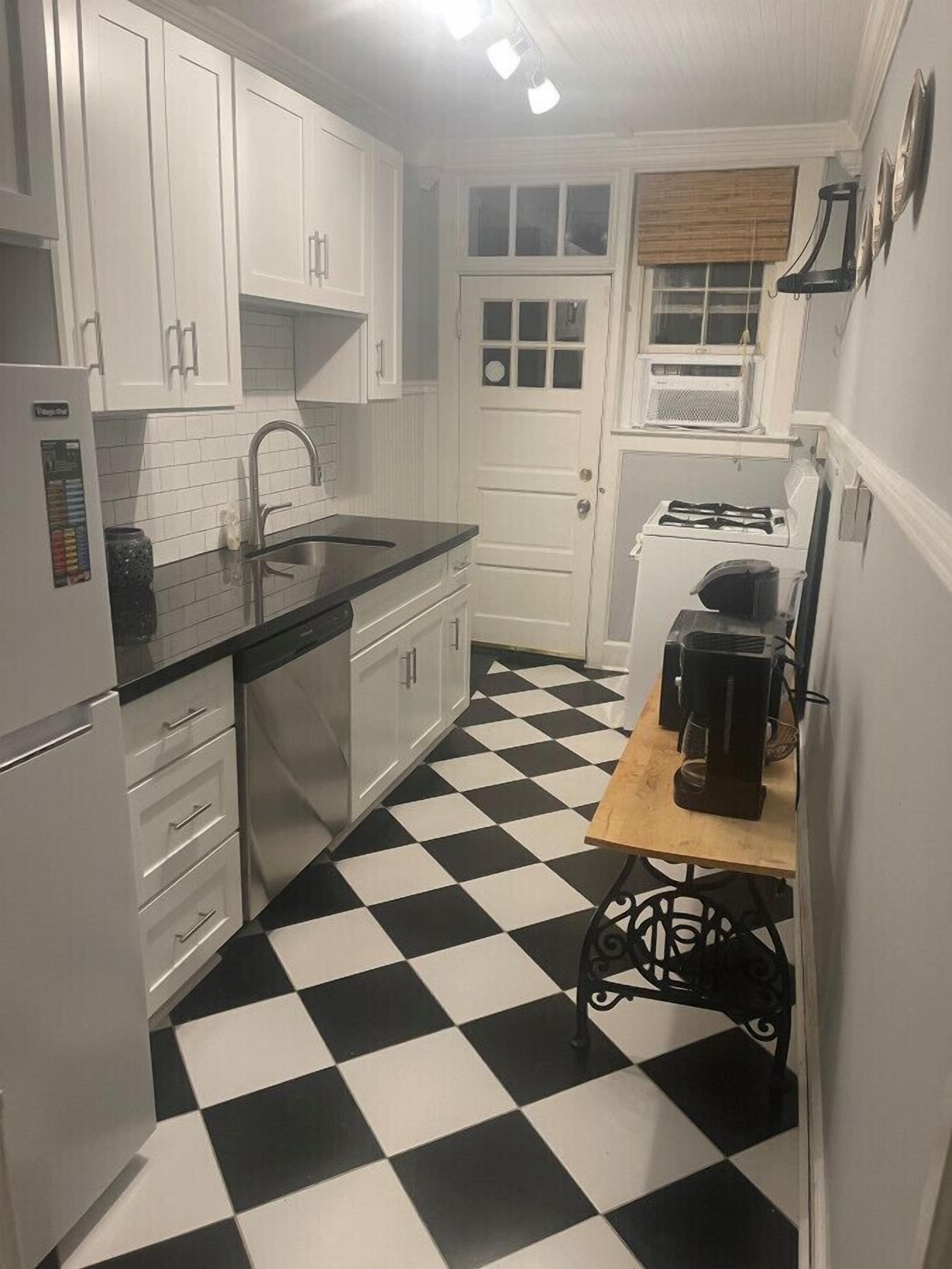 Una cucina compatta con mobili bianchi, pavimento a scacchi bianconero, un lavandino, un fornello e una macchina da caffè.