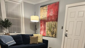 A living room with a dark blue sofa, patterned armchair, and a glass coffee table. A large abstract painting and a floor lamp are on the wall. - Fisher Park condo on Elm St., new kitchen, 1 mile from downtown (Greensboro)