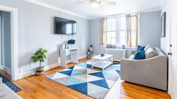 A modern living room with a grey sofa, a coffee table, a TV mounted on the wall, and a large window with curtains.