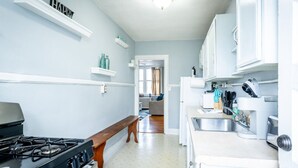 A kitchen with a stove, sink, and white cabinets. There is a bench in the middle of the room and a doorway leading to another room.