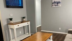 Una sala de estar con una mesa de centro de madera, una consola blanca con plantas, un sofá gris y un tapete estampado.
