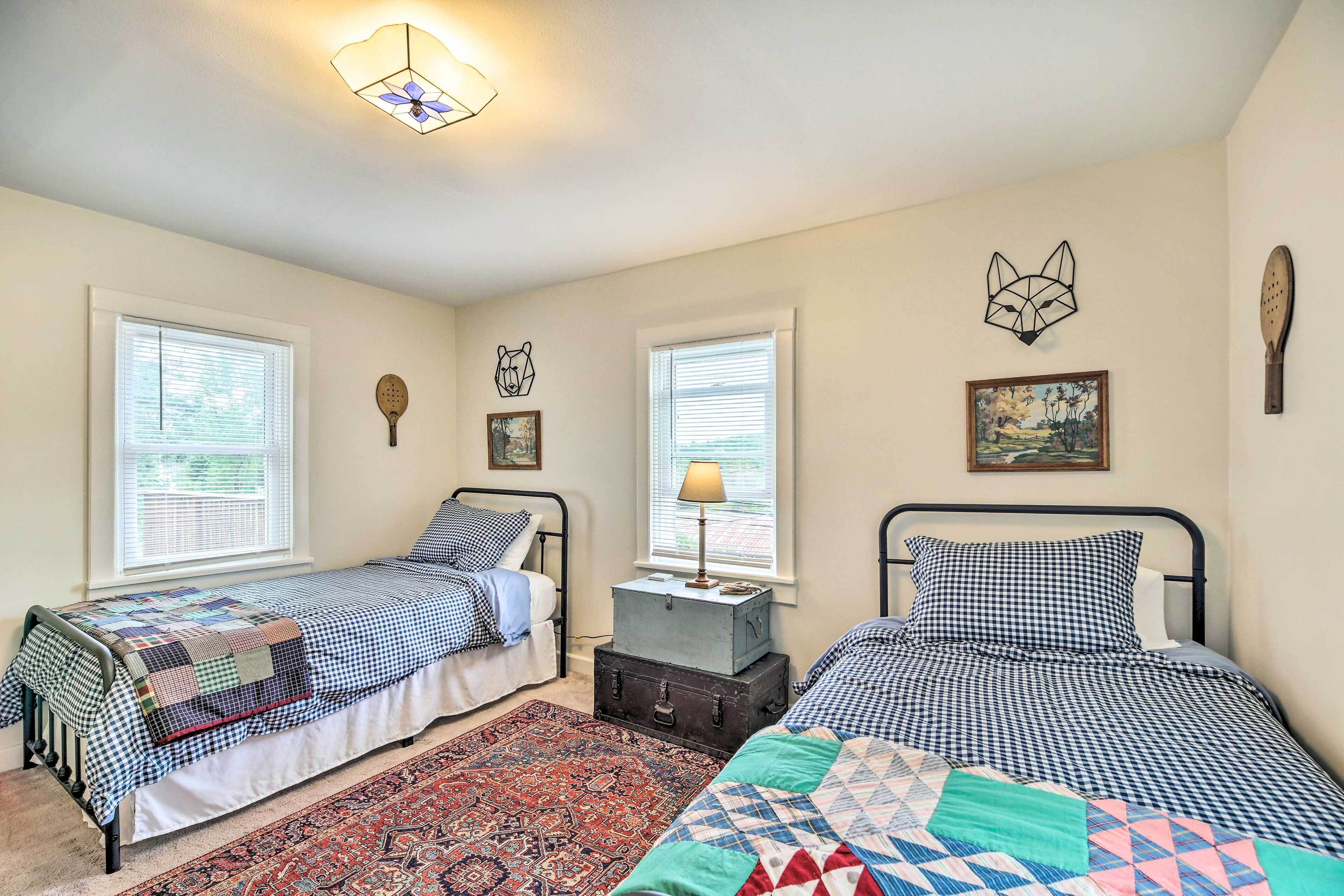 A bedroom with two single beds, a nightstand with a lamp, a wall clock, and a window with blinds.