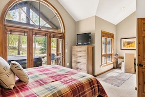 A bedroom with a large bed, a television, a desk, and a window overlooking a garden.