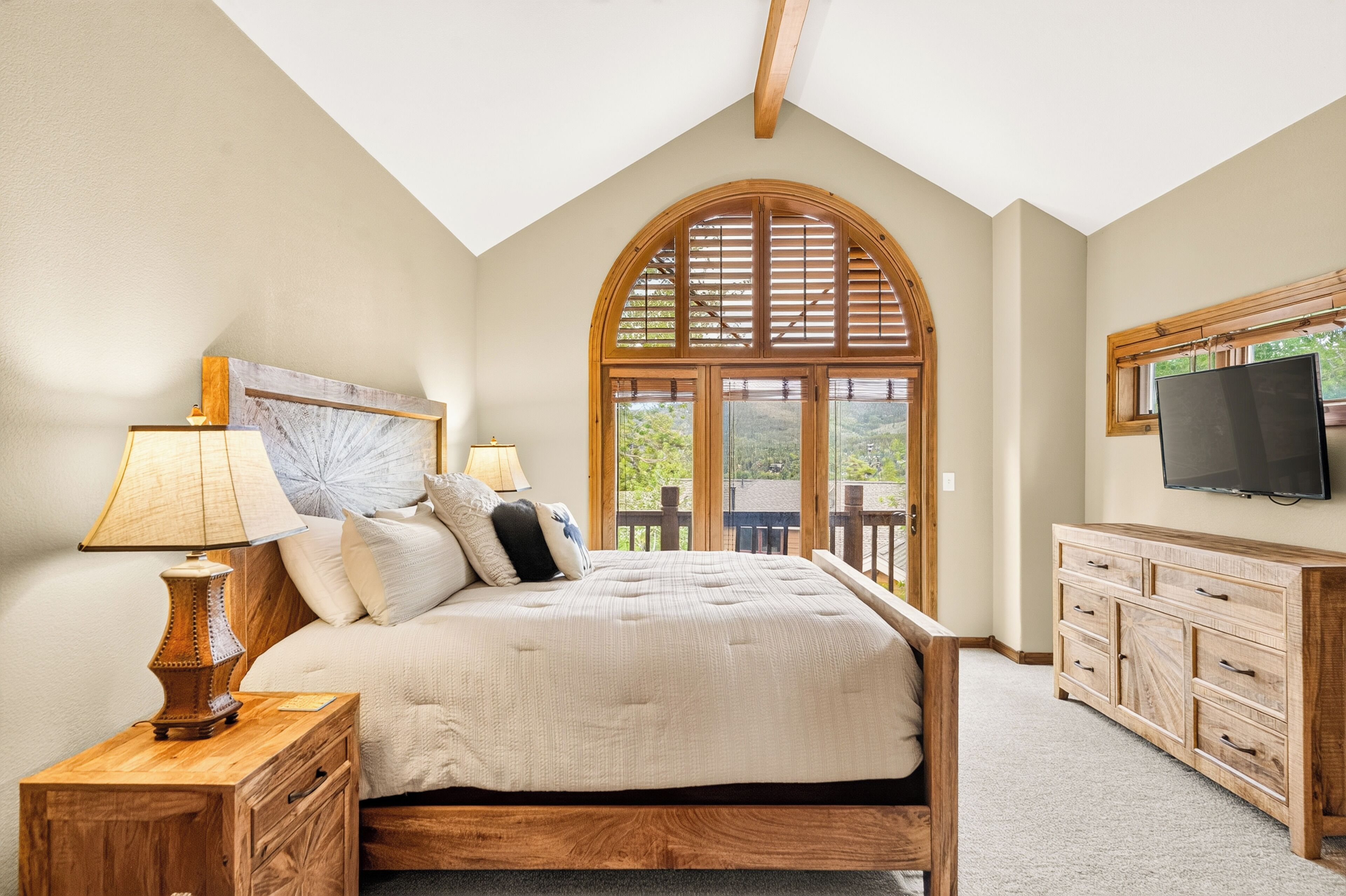 Ein Schlafzimmer mit einem großen Bett, einem Holzkopfstück, einem an der Wand montierten Fernseher und Blick ins Freie durch ein Fenster.