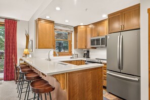 A modern kitchen with wooden cabinets, a stainless steel refrigerator, and a bar area with stools. - Close to Peak 9, On Main St, Shared Pool/Hot Tubs. (Breckenridge)
