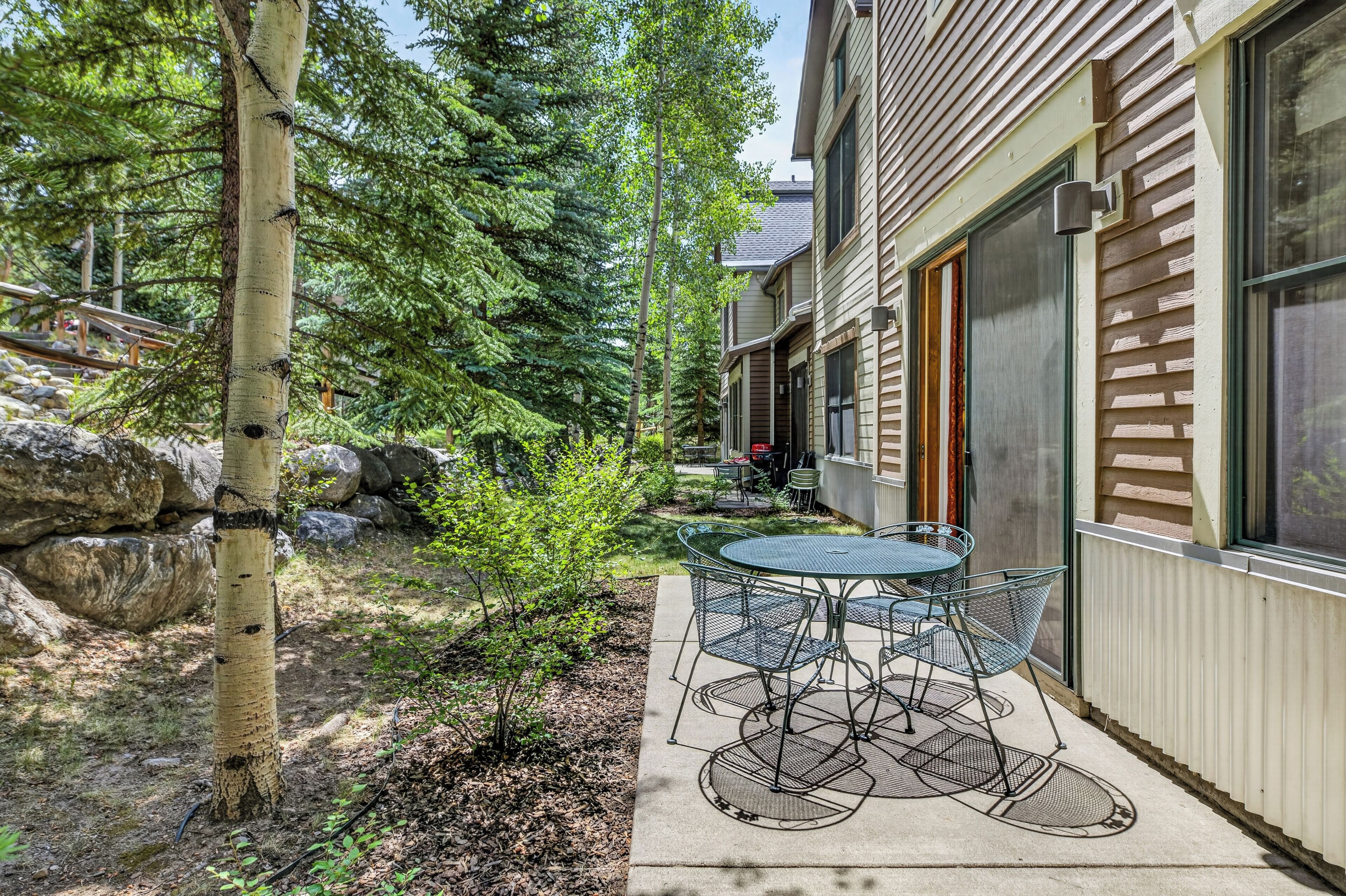 A residential property with a patio area featuring a table and chairs, surrounded by trees and a stone wall.