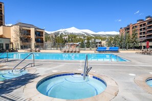 A pool area with multiple hot tubs and a large swimming pool, surrounded by buildings and a snow-capped mountain in the background. - Close to Peak 9, On Main St, Shared Pool/Hot Tubs. (Breckenridge)