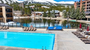 Une piscine entourée de bâtiments, avec des chaises longues et des parasols, donnant sur un lac et des montagnes en arrière-plan.