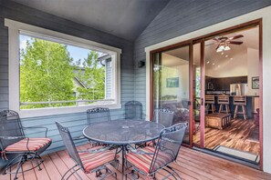 A covered patio with a round table and chairs, a sliding glass door to a kitchen, and a view of trees outside.