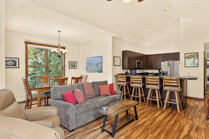 A modern living room with a grey sofa, wooden chairs, a dining table, and a kitchen with dark cabinets and stainless steel appliances.
