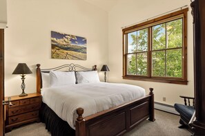 A bedroom with a wooden bed, two bedside lamps, a nightstand with drawers, a large window with a view of trees, and a landscape painting on the wall.