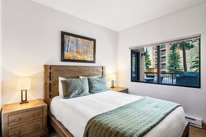 Une chambre d’hôtel avec un lit, deux tables de chevet, une fenêtre donnant sur des immeubles et des arbres, et un tableau accroché au mur.