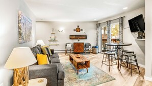 Een moderne woonkamer met een grijze bank, een houten salontafel en een eethoek met een ronde tafel en barstoelen.