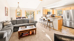 A modern living room with a gray sectional sofa, a wooden coffee table, and a dining area with bar stools. A kitchen with wooden cabinets and stainless steel appliances is visible.