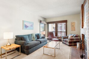Ein Wohnzimmer mit Sofa, Sesseln und Couchtisch. Ein Kamin mit Backsteinwand und ein Fenster mit Jalousien sind ebenfalls vorhanden.