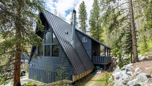 A modern cabin with a sloped roof, large windows, and a chimney. It is surrounded by trees and rocks, indicating a forested location. - Adventure Awaits! Cozy Aframe 5 Min to Breck. (Breckenridge)