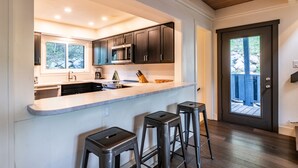 A modern kitchen with dark cabinets, a large island with stools, and a view through a door to an outdoor deck. - Adventure Awaits! Cozy Aframe 5 Min to Breck. (Breckenridge)
