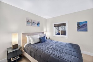 A bedroom with a bed, a nightstand, a bedside table, a window with a view, and wall art.