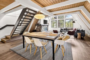 A spacious loft with a high ceiling, a wooden staircase, a dining area with a table and chairs, a living room with a sofa and a fireplace, and large windows offering a view of the outdoors. - Adventure Awaits! Cozy Aframe 5 Min to Breck. (Breckenridge)