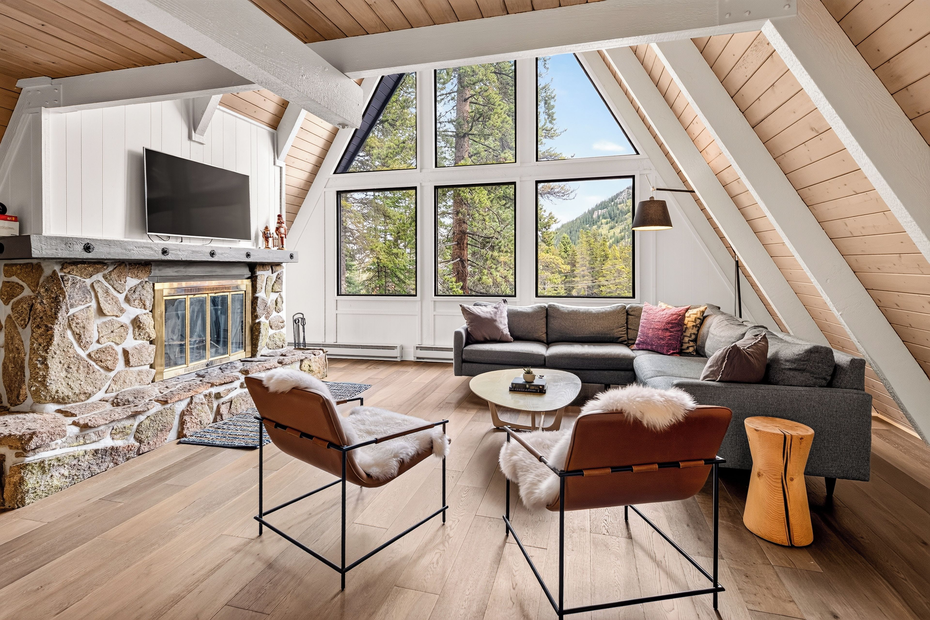 A modern loft with a stone fireplace, a large window with a view of trees, and a cozy seating area with leather chairs.