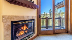 A fireplace with a visible fire, a wooden door with glass panels, and a view of trees and a wooden deck outside.