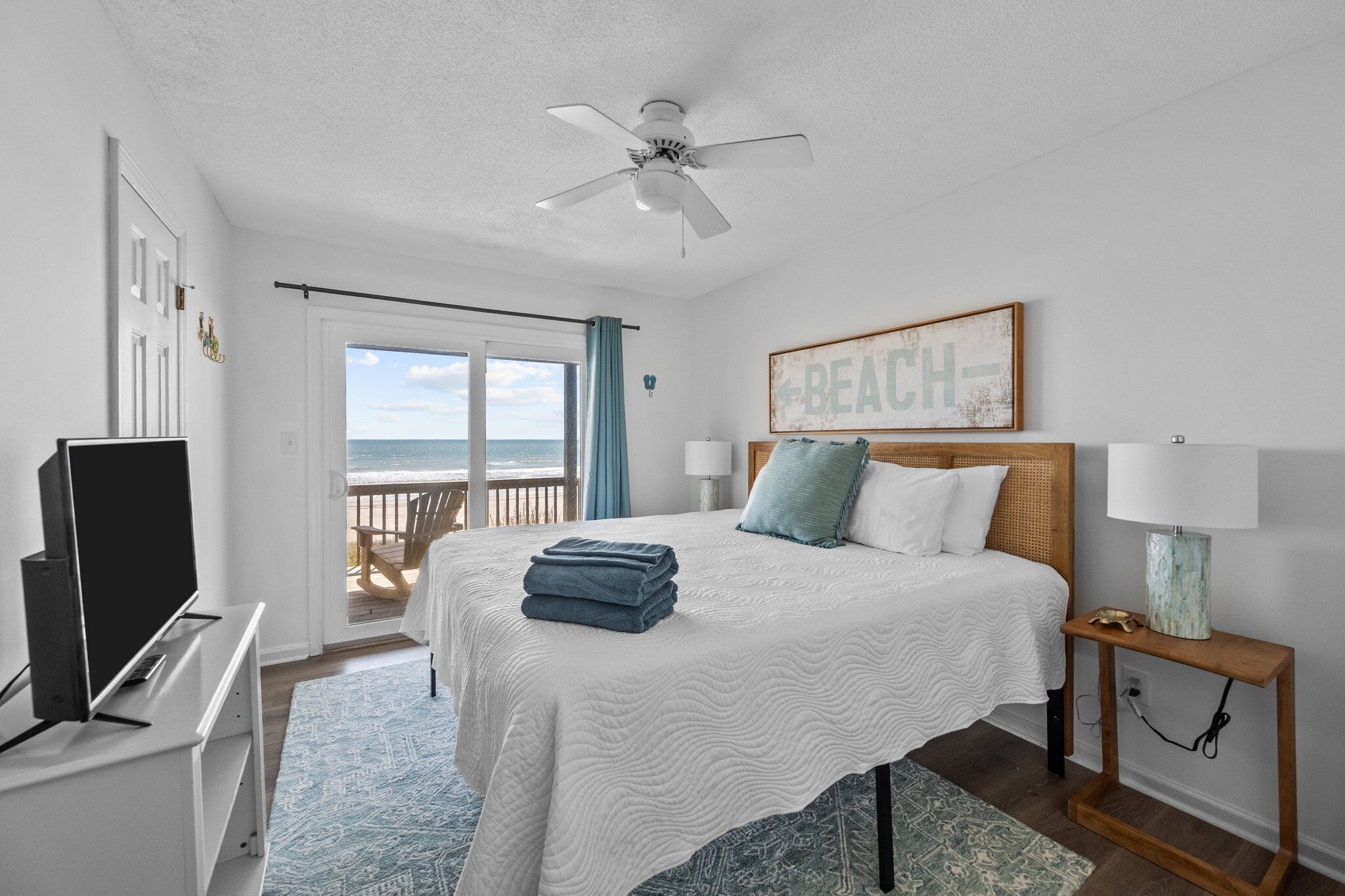 Ein Schlafzimmer mit Bett, Deckenventilator, Fernseher, Nachttisch und Blick auf den Strand durch eine Schiebetür.