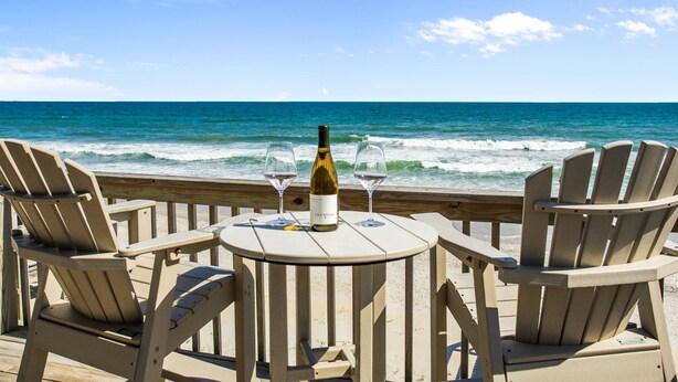 A wooden deck with four Adirondack chairs and a round table with a bottle of wine and two glasses. The deck overlooks the ocean with waves crashing onto the shore. - Oceanfront 4BR | Sandcastles + Golden Sunrises (Surf City)