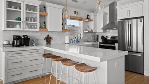 A modern kitchen with white cabinetry, stainless steel appliances, and wooden bar stools. - Oceanfront 4BR | Sandcastles + Golden Sunrises (Surf City)