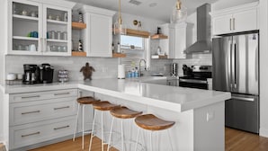 A modern kitchen with white cabinetry, stainless steel appliances, and wooden bar stools. - Twin Landings | Two Homes, One Epic Family Escape! (Surf City)