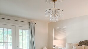 A bedroom with a large bed, a chandelier, a window with blinds, and a door with curtains. - 30a Calm Gulf Cottage -1 mile from beach + golf cart (Santa Rosa Beach)