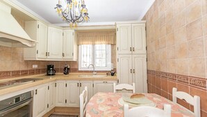 A kitchen with white cabinets, a window with a beige curtain, a chandelier, and a table with a floral tablecloth. - Casa La Mirada - RCP152 Cómpeta by Ruralidays (Málaga)