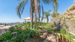 A garden with a pool, palm trees, and a stone wall. - Casa La Mirada - RCP152 Cómpeta by Ruralidays (Málaga)