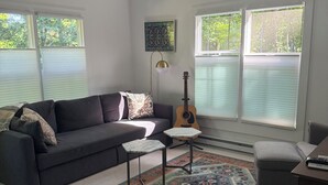 Un salon avec un canapé foncé, deux petites tables, une guitare, un tapis à motifs et de grandes fenêtres.