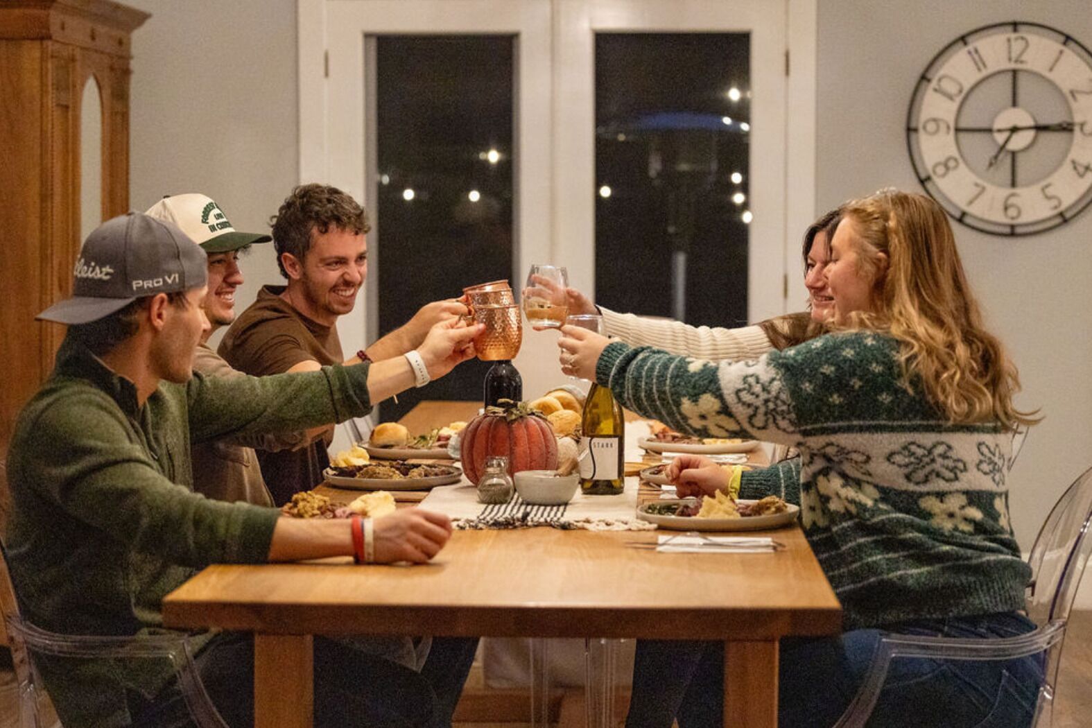 Een groep mensen die aanroept tijdens een diner met een pompoen als decoratie, een fles wijn en een klok aan de muur.