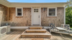 Ein Haus mit Veranda, einer weißen Tür, zwei Fenstern, einer kleinen Sitzgruppe im Freien mit zwei Stühlen und einer Holzterrasse.
