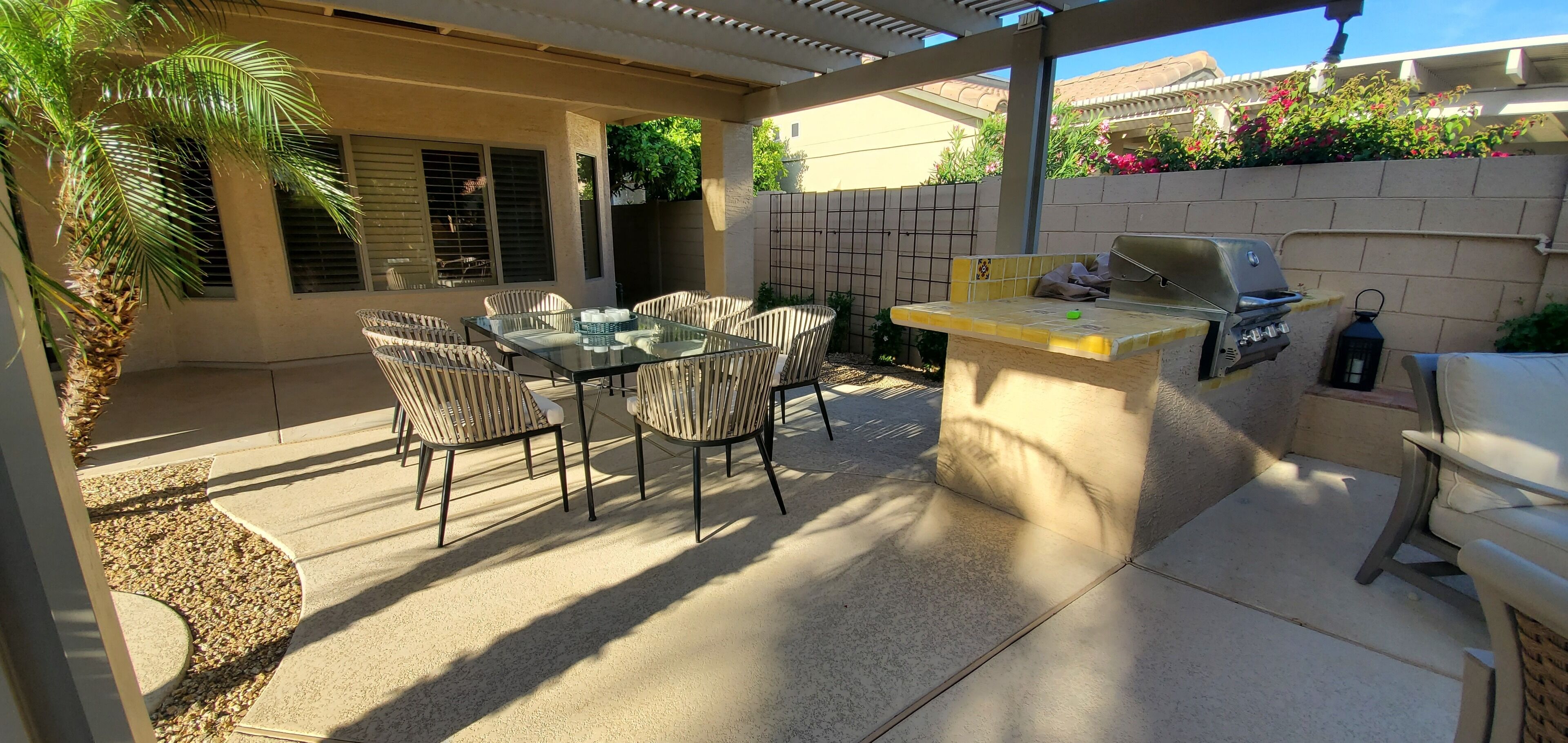 Een terras buiten met een glazen tafel, rieten stoelen, een barbecue en een palmboom.