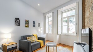 Una sala de estar moderna con un sofá gris, una mesita redonda pequeña, una lámpara blanca y un televisor sobre un mueble blanco.