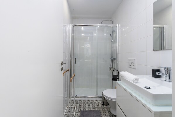 Un baño moderno con ducha envuelta en vidrio, inodoro blanco y lavamanos con superficie de vidrio.