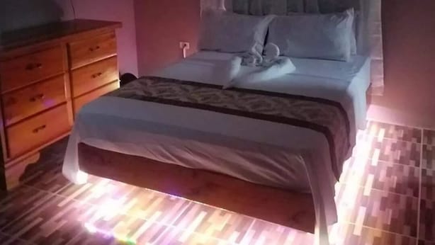 A bedroom with a bed, a wooden dresser, and a wall-mounted light fixture. - Mesmerize Guest House (Port Antonio)