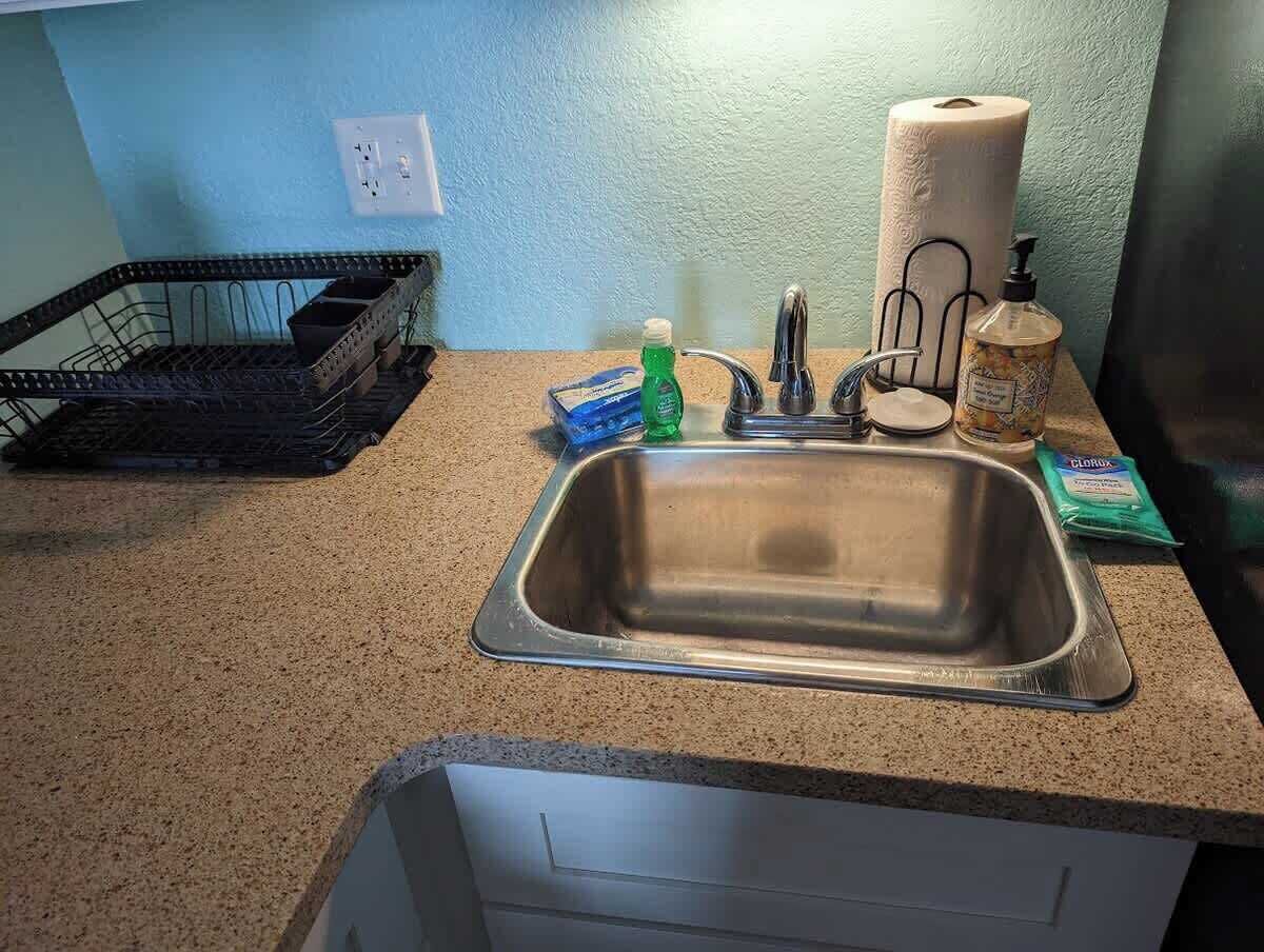 Una cocina con un lavamanos de acero inoxidable, una cesta de alambre negra, un rollo de toallas de papel, un dispensador de jabón y una botella de detergente para lavar platos.