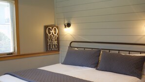 Un letto rifatto con ordine, struttura in metallo, luce fissata al muro e cartello incorniciato.