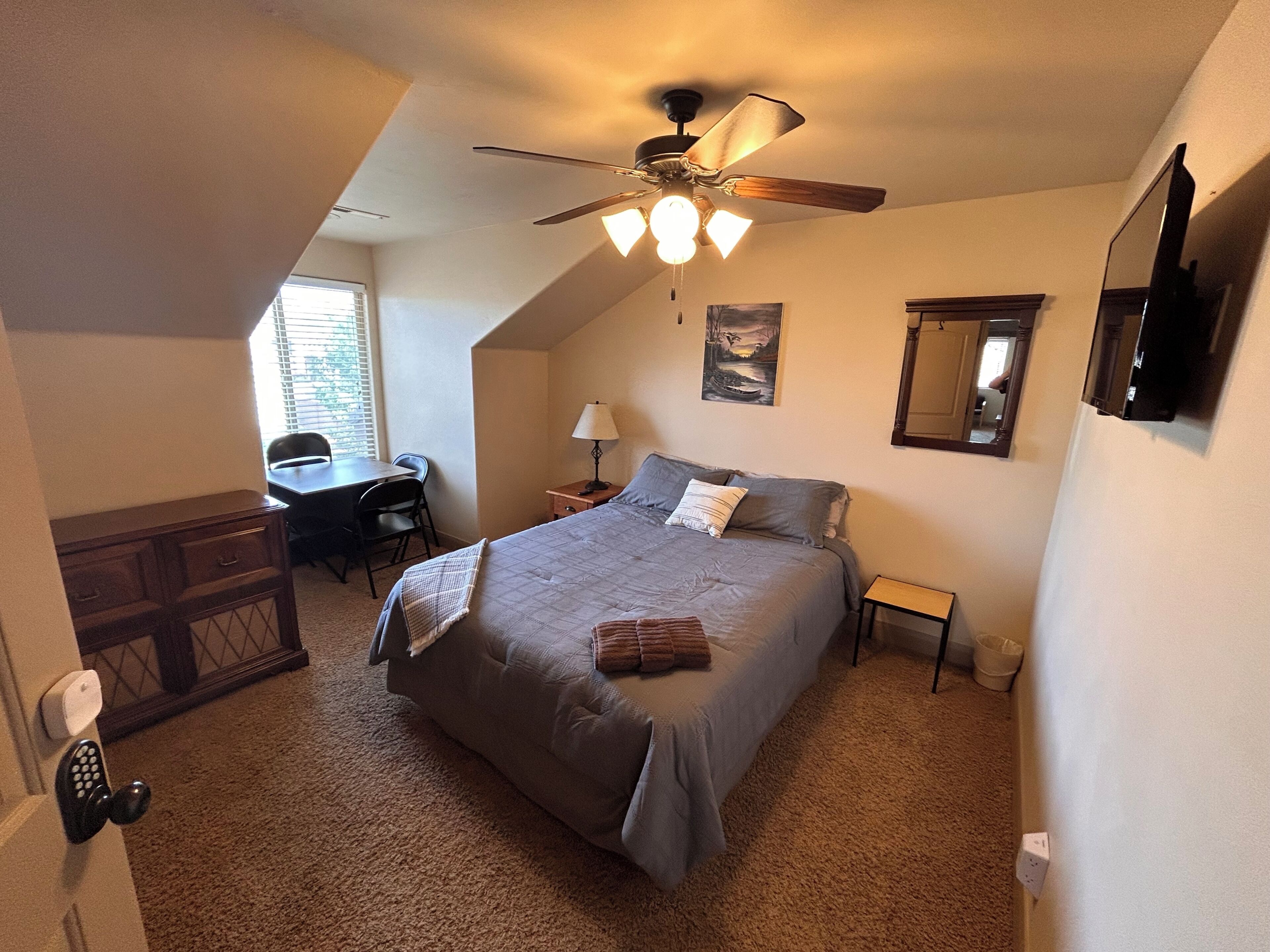 Une chambre à coucher comprenant un lit, un bureau, une chaise, un ventilateur de plafond, un tableau et un miroir.