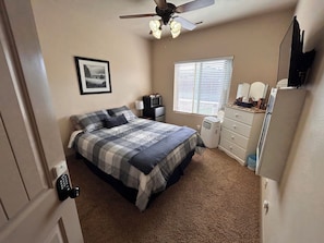 Une chambre à coucher comprenant un lit, un ventilateur de plafond, une fenêtre avec des stores, une commode et un four à micro-ondes.