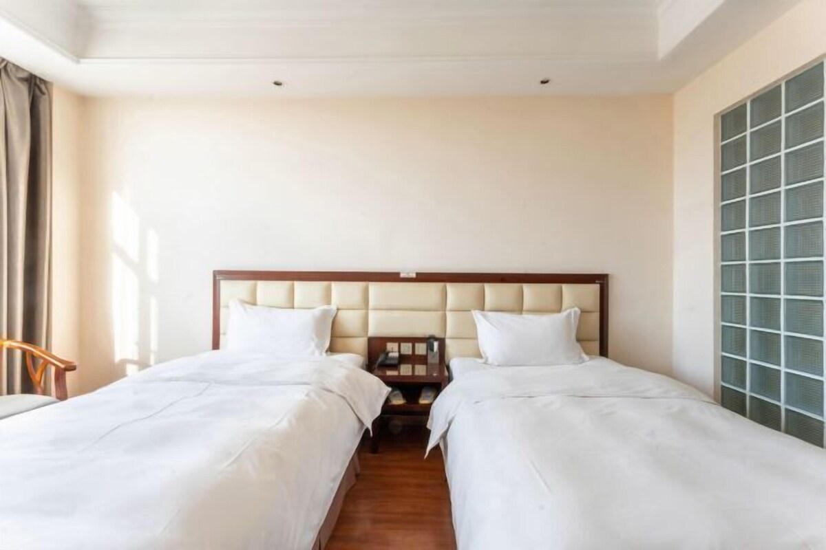 Ein Hotelzimmer mit zwei Einzelbetten, einem Holzkopfteil, einem Nachttisch und einem großen Fenster mit Jalousien.