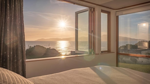 A bedroom with a large window overlooking a scenic view of the sea and distant mountains during sunset. - Seascapes Beach House (Paraparaumu)