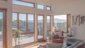 A spacious room with large windows offering a view of a wooden deck and mountains. - Seascapes Beach House (Paraparaumu)