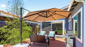 Un patio esterno con tavolo e sedie, un ombrellone grande e un barbecue.